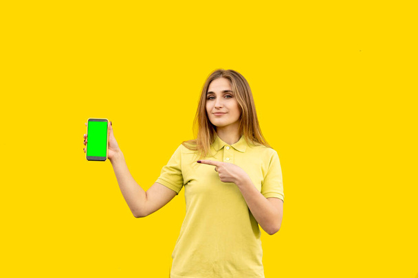 Genç mutlu kız telefonu işaret ediyor. Başvurusunun ne olduğunu gösteriyor. Sarı bir tişört giymişti. Sarı arkaplanda izole edilmiş. - Fotoğraf, Görsel