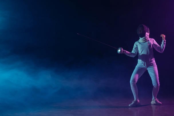 Schwertkämpferin in Fechtanzug und Maske hält Degen auf schwarzem Hintergrund mit Rauch und Beleuchtung  - Foto, Bild