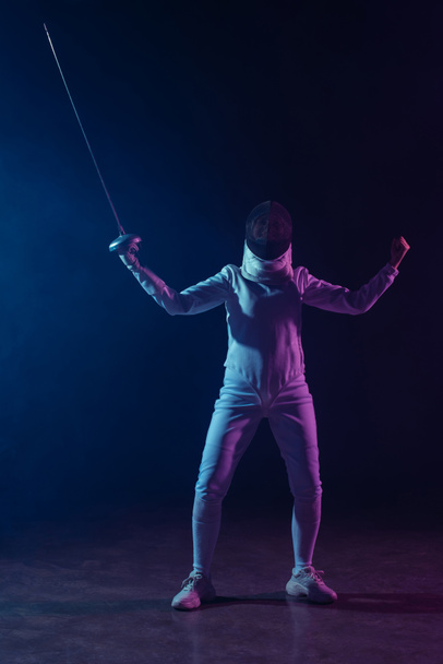 Fencer showing yeah gesture while holding rapier on black background with lighting  - Foto, immagini