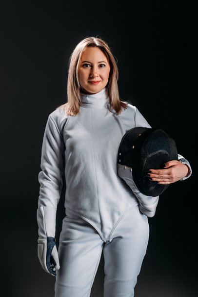 Beautiful fencer looking at camera while holding fencing mask isolated on black  - Photo, Image