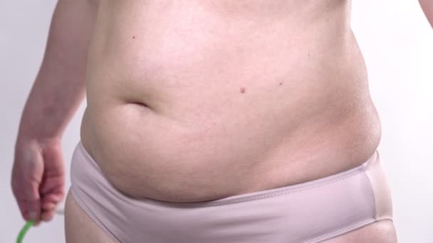 Une femme d'âge moyen à la peau affaissée définit son gros ventre avec un ruban à mesurer, sur un fond blanc. Plan latéral. - Séquence, vidéo
