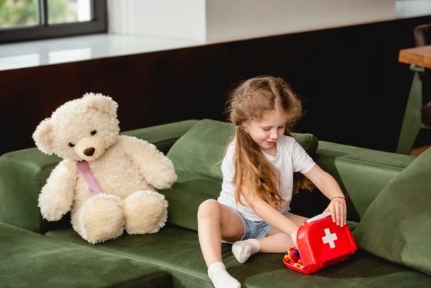 cute child touching toy first aid kit box near teddy bear while playing doctor game  - Foto, Imagem