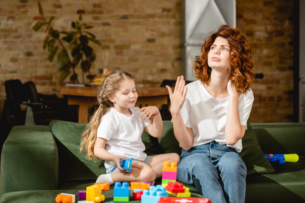 redhead mother feeling hot and waving hand near adorable daughter and building blocks  - Photo, Image