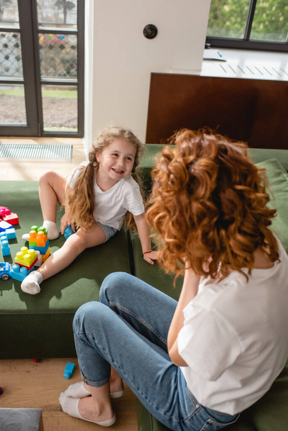 Selektive Fokussierung des fröhlichen Kindes auf die rothaarige Mutter in der Nähe von Bauklötzen auf dem Sofa - Foto, Bild