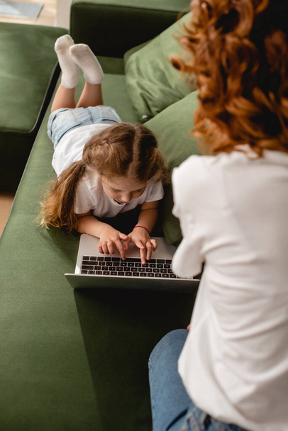 back view of curly mother near daughter using laptop in living room  - Photo, Image