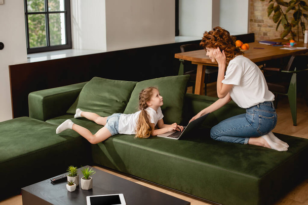 cute kid using laptop near curly mother in living room  - Foto, afbeelding