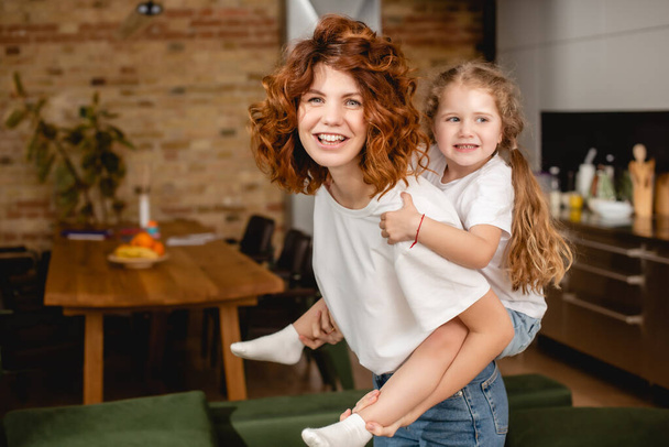 cheerful and curly mother piggybacking cute daughter  - Zdjęcie, obraz