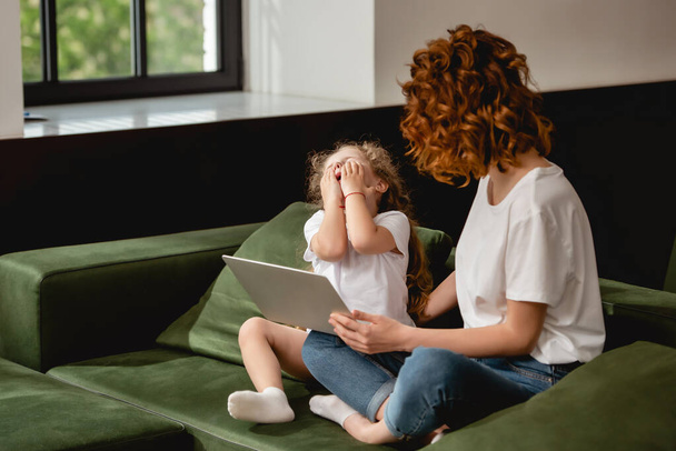 madre rizada sosteniendo portátil cerca de hija emocional en la sala de estar
   - Foto, Imagen