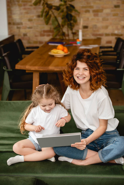 веселая рыжая мать держит ноутбук рядом с милой дочерью в гостиной
  - Фото, изображение