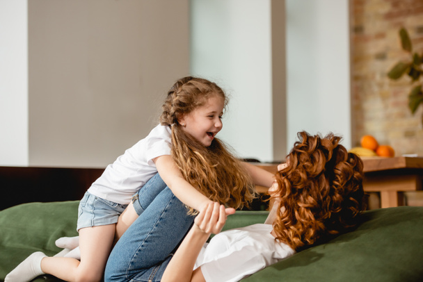 curly mother lying on sofa, lifting cheerful child and holding hands in living room  - Photo, Image