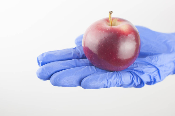 Рука в синіх нітрилових рукавичках тримає свіже червоне яблуко на білому
. - Фото, зображення