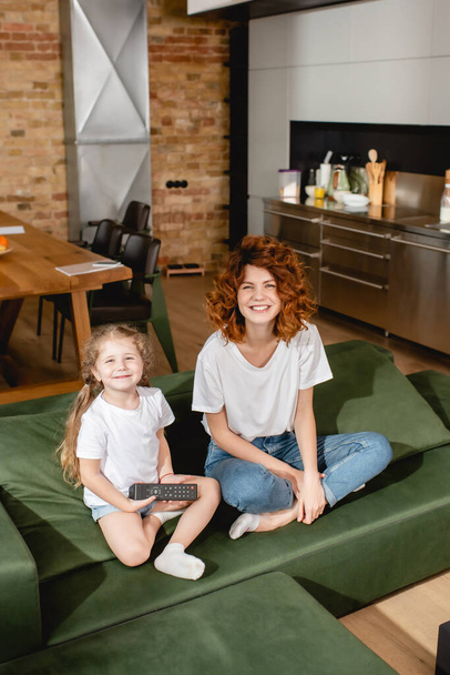 cute kid holding remote controller and sitting with redhead mother on sofa - Fotoğraf, Görsel