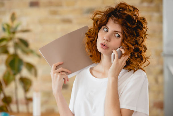 surprised redhead woman holding folder and talking on smartphone  - Foto, Imagen
