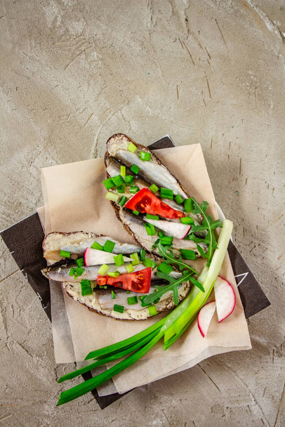 heerlijke sandwiches met brood, boter, kleine sardines, plakjes tomaten, radijs en groene uien op voedselperkament op een betonnen achtergrond, ongewone stijlvolle presentatie van de schotel, witte ruimte - Foto, afbeelding