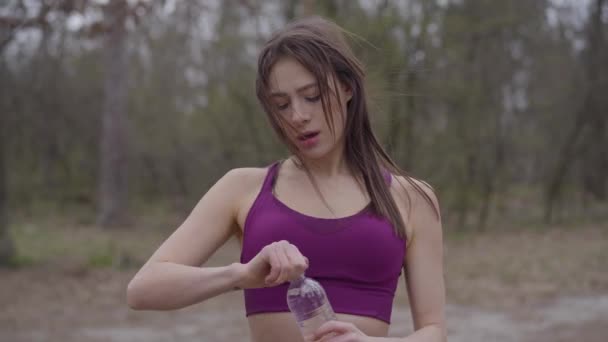 Portrait of young beautiful Caucasian woman with heart tattoo on hand drinking water during workout outdoors. Tired sportswoman having break in training in the park. Fitness, sport, athleticism. - Záběry, video