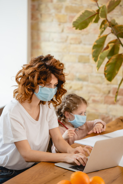 selective focus of curly freelancer mother in medical mask using laptop near daughter at home - Foto, Bild