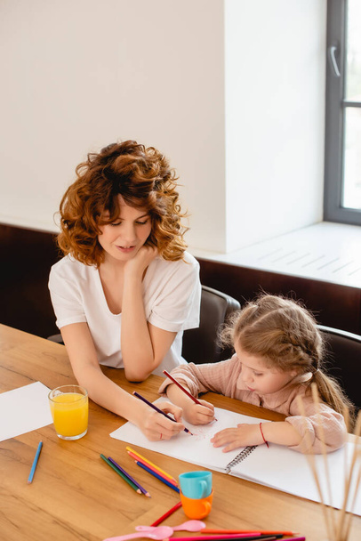 curly mother drawing with cute daughter at home - Фото, изображение