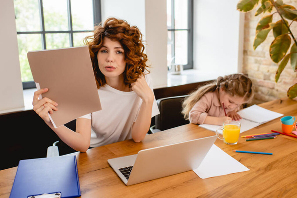 curly freelancer mother holding folder near laptop and daughter drawing at home - Foto, Imagem