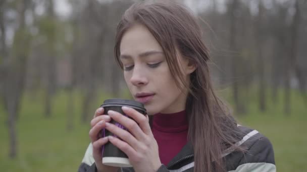 Close-up of thoughtful charming girl drinking coffee outdoors. Portrait of beautiful brunette woman with brown eyes enjoying cloudy day in park. Lifestyle, leisure, resting, nature. - Кадри, відео