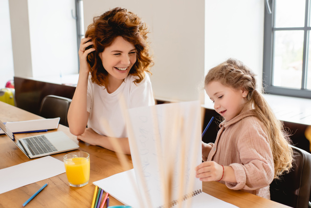 selective focus of curly mother smiling near kid and laptop  - Фото, зображення