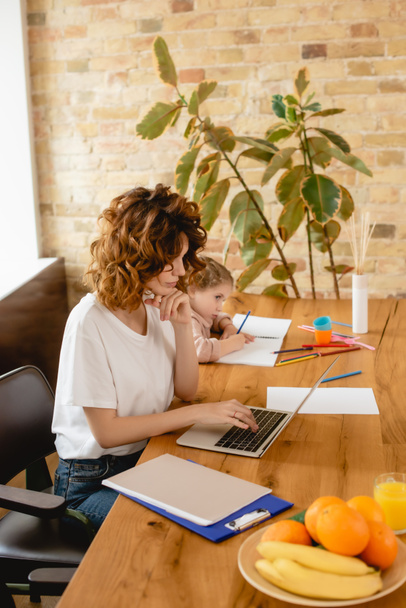 szabadúszó anya használ laptop, míg aranyos lánya rajz otthon - Fotó, kép