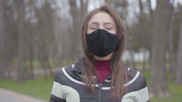 Confident young Caucasian woman in face mask looking around in empty park. Brunette girl standing outdoors during Covid-19 quarantine. Coronavirus pandemic, infection outbreak, lifestyle. - Felvétel, videó