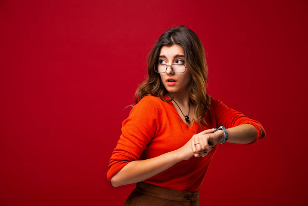 The girl in glasses looks at the watch. Hurry up. Appreciate your time. Do not be late. Isolated on red background. - Photo, image