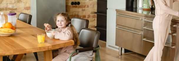 orientação panorâmica da criança feliz perto da tigela com flocos de milho, vidro de suco de laranja e mãe
  - Foto, Imagem