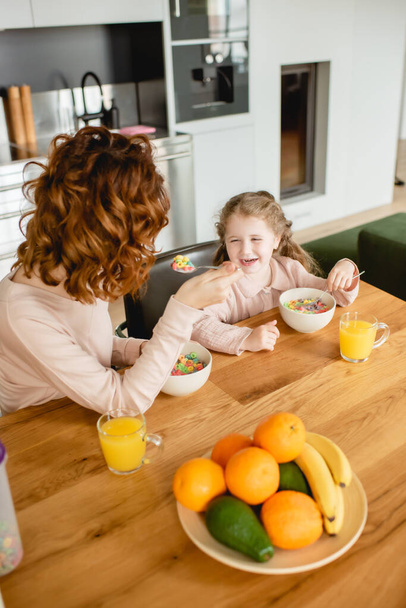 madre rizada y feliz hija sosteniendo cucharas cerca de cuencos con hojuelas de maíz y vasos de jugo de naranja
  - Foto, imagen