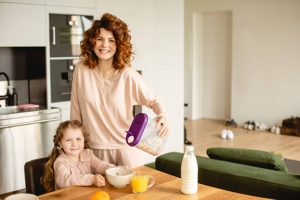 glückliche Mutter hält Behälter mit Cornflakes in der Nähe der Tochter, Flasche mit Milch und einem Glas Orangensaft  - Foto, Bild