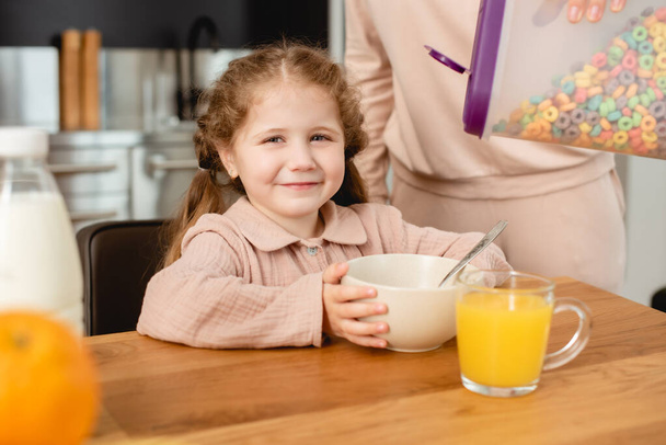 selective focus of mother holding container with delicious corn flakes near bowl and cute daughter  - Photo, Image