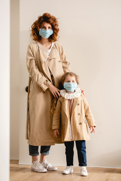 curly mother and cute daughter in medical masks standing in trench coats  - Foto, Bild