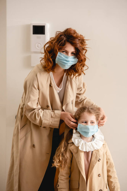 σγουρή κοκκινομάλλα μητέρα με καμπαρντίνα φορώντας ιατρική μάσκα σε χαριτωμένη κόρη  - Φωτογραφία, εικόνα