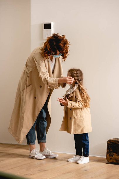 curly mother in trench coat wearing medical mask on kid  - Фото, изображение