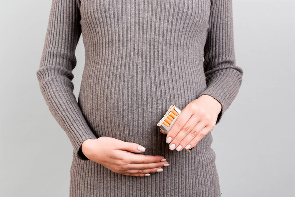 グレーの背景に妊婦の手にタバコのパックを閉じます。タバコとニコチン中毒。赤ん坊は危険にさらされている. - 写真・画像