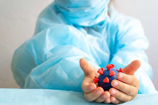 Kind in medizinischem Gewand und Schutzmaske hält in seinen Händen ein Modell des Coronavirus aus Knetmasse auf weißem Hintergrund. Isoliert auf weiß. Kopierraum. Gesundheitswesen, Medizin, Covid-19 - Foto, Bild
