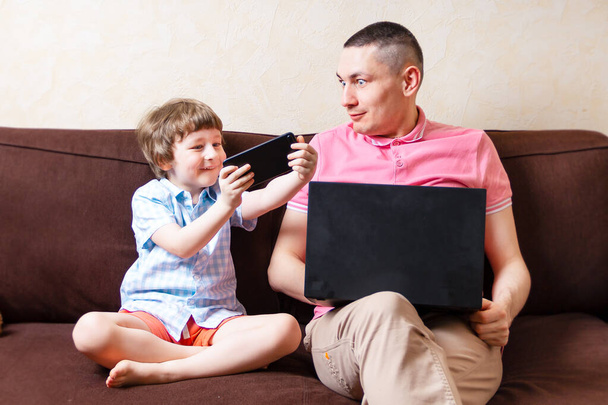 驚きの若い男は、幼児の息子とノートパソコンのリラックスを使用してコンピュータ上で一緒にお楽しみください座って、お父さんと小さな男の子の子供はガジェットで忙しいソファの上で自宅で休息でスマートフォンをお楽しみください。オンライン教育 - 写真・画像