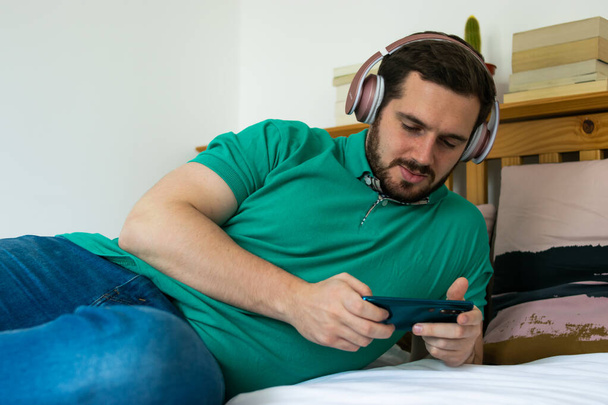 Photo d'un jeune et séduisant homme barbu assis sur le lit avec des écouteurs roses jouant avec un jeu sur ce téléphone d'une manière relaxante
. - Photo, image
