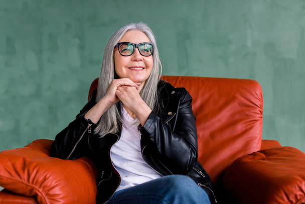 Beautiful stylish senior woman with long straight gray hair, wearing eyeglasses and black leather jacket, sitting in comfortable sofr red armchair on green background and dreamly looking aside - Фото, изображение