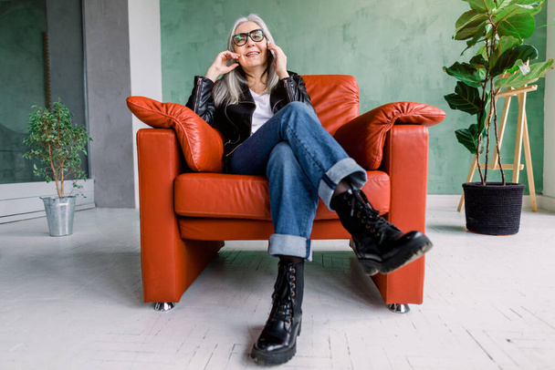 Full length portrait of attractive smiling modern senior woman with long staright gray hair, wearing trendy leather jacket and jeans, sitting in comfortable chair and talking phone with friend - Фото, изображение