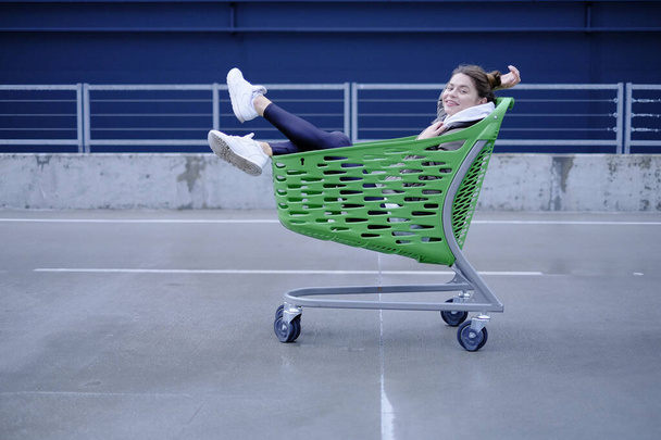 A slender dark-haired lady, dressed in casual attire, is having fun with a grocery cart near the shopping center. - Φωτογραφία, εικόνα