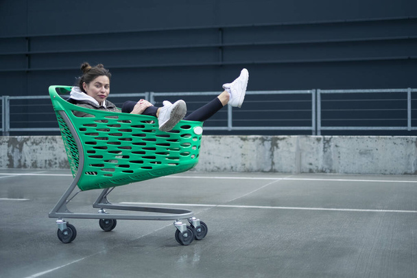 A slender dark-haired lady, dressed in casual attire, is having fun with a grocery cart near the shopping center. - Foto, afbeelding