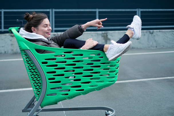 A slender dark-haired lady, dressed in casual attire, is having fun with a grocery cart near the shopping center. - Zdjęcie, obraz