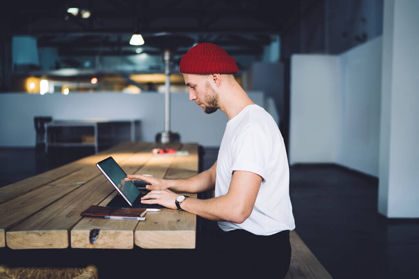 Side view of focused professional casual hipster adult worker in red beany sitting at wooden desk using notebook with touchscreen at loft office - Zdjęcie, obraz