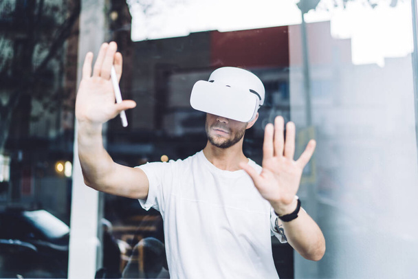 Young immersive bearded male graphic designer in white shirt wearing virtual reality headset testing app using hands and pen behind glass - Foto, Imagen