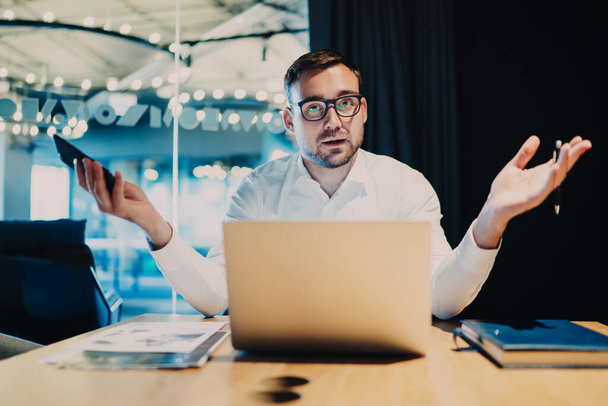 Surprised man in formal wear and glasses working on startup presenting business meeting gesturing sitting at table in conference hall - Foto, Bild