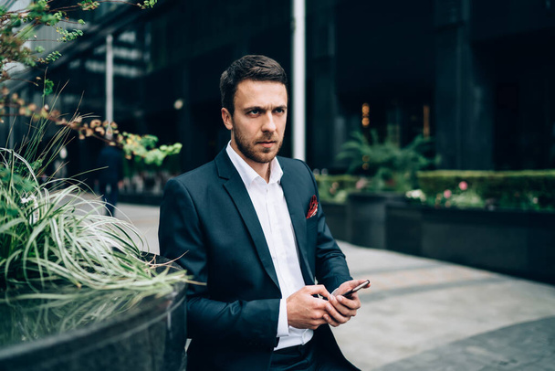 Pensive adult male in formal wear thinking and looking away while sitting near flowerbed and using smartphone in downtown of modern city - Фото, изображение