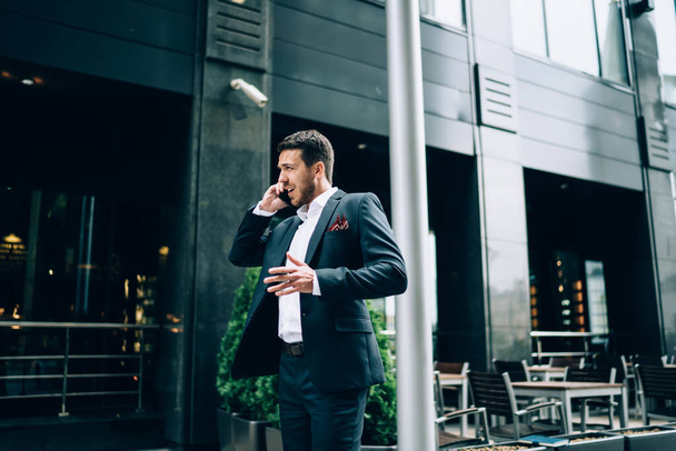 Furious adult businessman in dark formal wear gesticulating and scolding worker during smartphone conversation outside modern building on city street - Photo, Image
