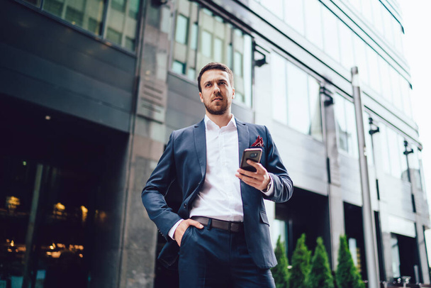 Low angle of frowning businessman with hand in pocket of formal wear looking away and browsing smartphone while standing on street of modern city - Foto, Bild