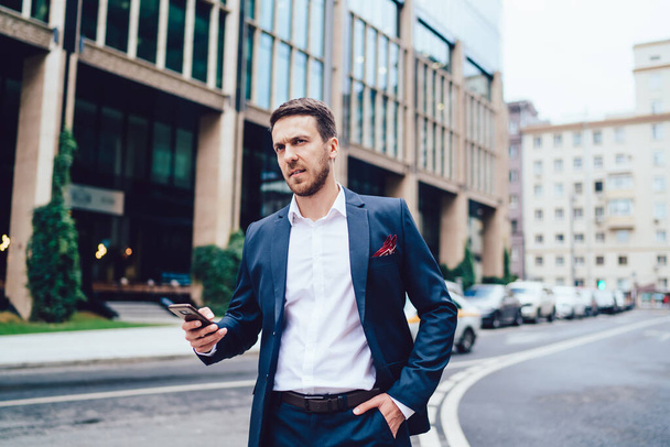 Serious boss with hand in pocket of formal wear looking away and frowning while standing near road and browsing smartphone in downtown - Fotografie, Obrázek